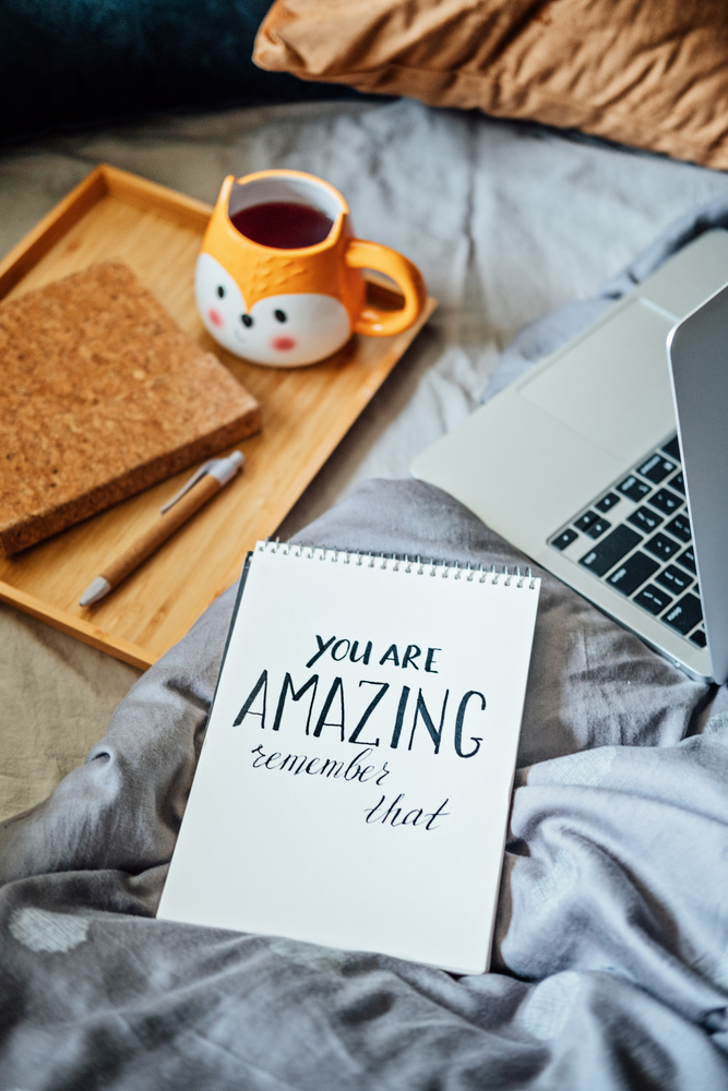 Positive daily affirmations for self love. Words You Are Amazing in notebook near laptop and tea coffee cup at bad in home. Handwritten affirmations text You Are Amazing in notepad